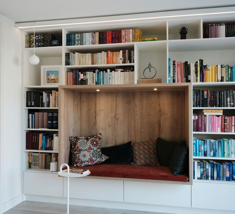 Boekenkast met geïntegreerde zithoek, houten panelen, kleurrijke kussens, moderne verlichting, witte afwerking, diverse boeken.